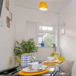 Dining area with a glass table set for four, yellow chairs, orange placemats, white walls, and a yellow pendant light.