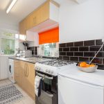 Modern kitchen with black subway tile backsplash, light wood cabinets, and an orange window shade over the sink area. A gas stove, toaster, and stainless appliances line the counter.