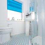 White bathroom with a blue roller blind over the window, toilet on the left, pedestal sink, and curved glass shower enclosure to the right.