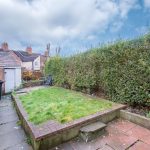 Residential backyard with a tall green hedge along the right, brick steps and patio, and a wooden fence on the left under a cloudy sky