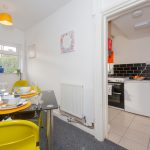 Bright dining area with a glass table set for four and yellow chairs, plants by a window, leading to a compact kitchen with a black tile backsplash.