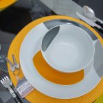 Close-up of an elegant place setting with an orange placemat, gray and white dishes, a spoon and knife, and a wine glass on a dark table.