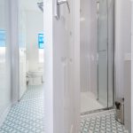 Narrow bathroom with blue-and-white geometric tile floor, open door showing a toilet room, and a glass shower enclosure on the right.