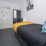 A small bedroom with a dark bed, yellow throw, and a colorful sunglasses-pattern pillow; a white wall and grey furniture including a wardrobe and chest of drawers, and a door on the left with a radiator nearby.