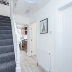 Bright hallway with grey carpet stairs on the left, white walls, and a doorway to a kitchen visible straight ahead; a framed picture on the wall and a door labeled '1' on the right.
