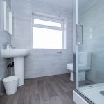 Modern grey-tiled bathroom with a glass shower enclosure, pedestal sink, toilet, and chrome towel radiator.