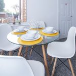 Round glass dining table set for four with white dishes on yellow placemats and white chairs in a bright room.