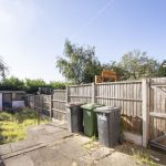 Backyard scene with a tall wooden fence, three wheeled rubbish bins (green and black) on a concrete patio, and overgrown grass nearby.