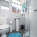 Small modern bathroom with a toilet, pedestal sink, glass shower, blue bath mat, and turquoise towels.