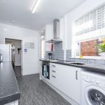 Bright modern kitchen with white cabinets, gray countertops, oven and stove, a washing machine under the counter, and a dish rack by the sink near a large window.