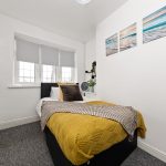 A small white bedroom with a bed dressed in a yellow throw and two pillows, next to a window with blind shades.