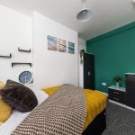 Modern bedroom with white walls and a green accent wall, bed with yellow pillows and brown patterned blanket, three-panel beach art above the headboard.