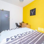 Bedroom with a bold yellow feature wall and a black deer head artwork, a desk and chair, and a tiled bedspread in the foreground.