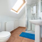 Bright bathroom with a glass shower enclosure, pedestal sink, white tiles, terracotta floor, and a skylight window in the sloped ceiling