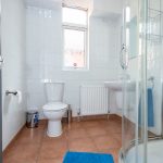 Bright white-tiled bathroom with a toilet, sink, radiator, and a curved glass shower; blue bath mats on terracotta floor. Regarded as a clean, modern bathroom design.