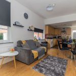 Open-plan living room with gray sofa, yellow throws, round side table, and a kitchen/dining area in the background.