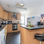 Bright modern kitchen with light wood cabinets, black countertops, an island with a bottle and glasses, and stainless steel appliances including oven and range hood.