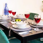 5f29953f-286c-4258-ac7c-7a350a32002djpg | RENT A ROOM | Professional House Shares in Warwickshire & West Midlands Dining table set for a meal with red wine glasses, white bowls, patterned placemats, and a blue bottle on a dark table; green chairs and a leafy plant in the background. Watermark RENTAROOM in corner.