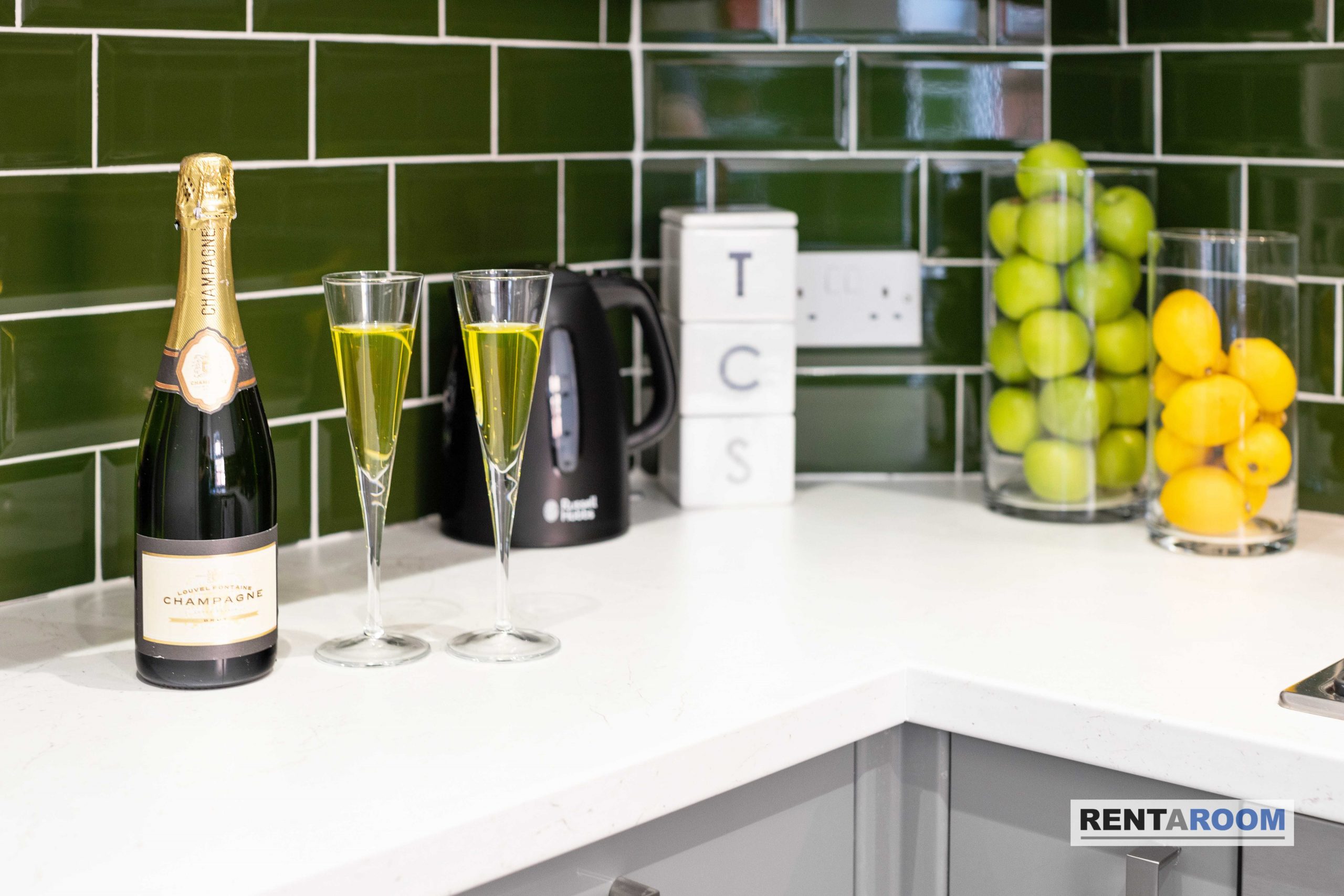 5f299540-2654-4eb7-9a32-79eb0a32002djpg | RENT A ROOM | Professional House Shares in Warwickshire & West Midlands Champagne bottle with two fluted glasses of bubbly on a white countertop, green tile backsplash in the background.