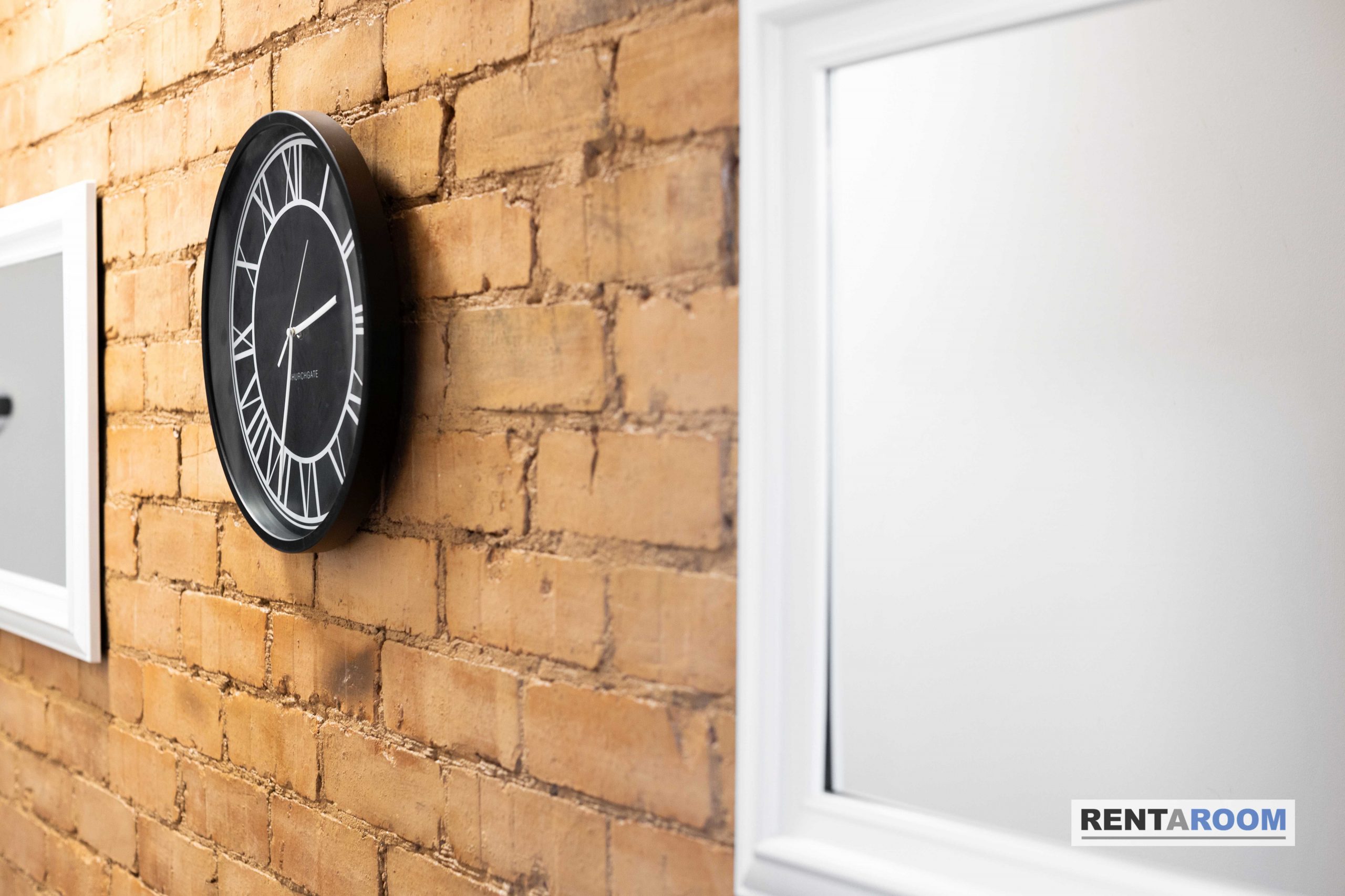 5f299541-da34-4034-9a03-7a350a32002djpg | RENT A ROOM | Professional House Shares in Warwickshire & West Midlands Round black clock with white Roman numerals mounted on an exposed brick wall beside a white door frame.