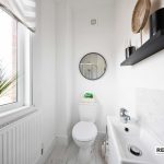 5f299542-90d4-4605-992d-7a350a32002djpg | RENT A ROOM | Professional House Shares in Warwickshire & West Midlands Bright white bathroom with a round wall mirror, sink, toilet, and a tall black shelf; large window with a green plant on the sill adds natural light.