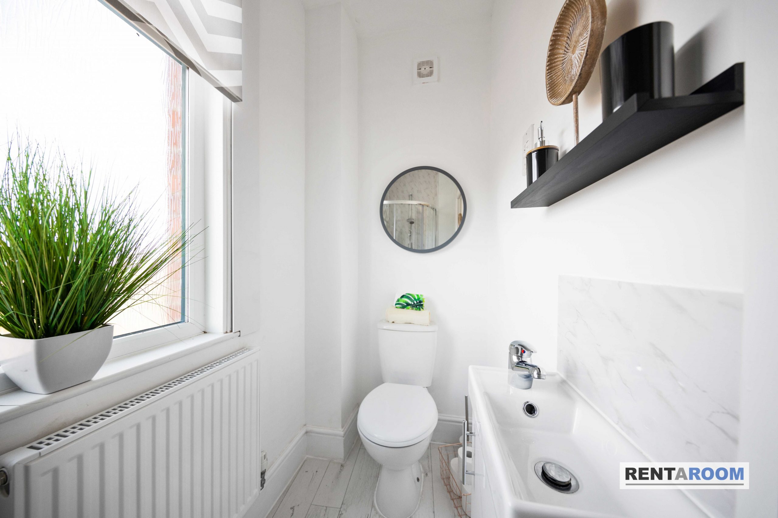 5f299542-90d4-4605-992d-7a350a32002djpg | RENT A ROOM | Professional House Shares in Warwickshire & West Midlands Bright white bathroom with a round wall mirror, sink, toilet, and a tall black shelf; large window with a green plant on the sill adds natural light.