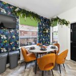 Round dining table set for six with orange chairs in a tropical floral room; window, TV and green blinds nearby.