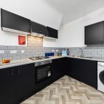 Modern black-cabinet kitchen with a gas stove, hood, and stainless appliances against a light gray tiled backsplash and white walls.