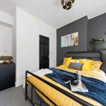 Modern bedroom with a black metal bed frame, yellow quilt, and blue throw against a dark gray accent wall with decor above the bed