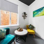 Cozy seating nook with teal and yellow chairs, a round table, plant shelf, and an abstract green-yellow wall art near a window showing a brick exterior.