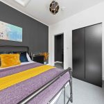 Modern bedroom with dark accent wall, metal bed frame, and colorful pillows (yellow, blue) on a purple throw bedspread.