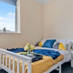 White wooden bed with a yellow quilt, blue throw, and geometric pillows in a sunlit bedroom near a window.