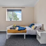 Bedroom with a white bed frame, mustard comforter, blue blanket, and striped pillows; window with a gray blind and trees outside.