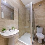 Modern beige marble bathroom with glass shower enclosure, white pedestal sink, toilet, and small potted plants on the sink. The space is clean and contemporary.