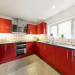 Modern red kitchen with glossy cabinets, white subway tile backsplash, stainless steel oven and range hood, and a large window with blinds.