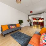 Bright open-plan living room with gray sofas, orange cushions, and a red-accent kitchen in the background.