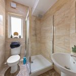Compact beige-tiled bathroom with a glass shower enclosure, a wall-mounted sink, and a toilet, under a slanted ceiling.
