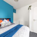 Bedroom with teal accent wall behind a bed adorned with blue, orange, and patterned pillows; white door and a hanging plant nearby.