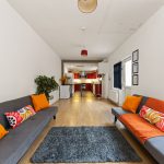 Bright open-plan living room with two long gray sofas facing each other, orange cushions, and a dark gray rug; kitchen with red cabinets visible in the background.