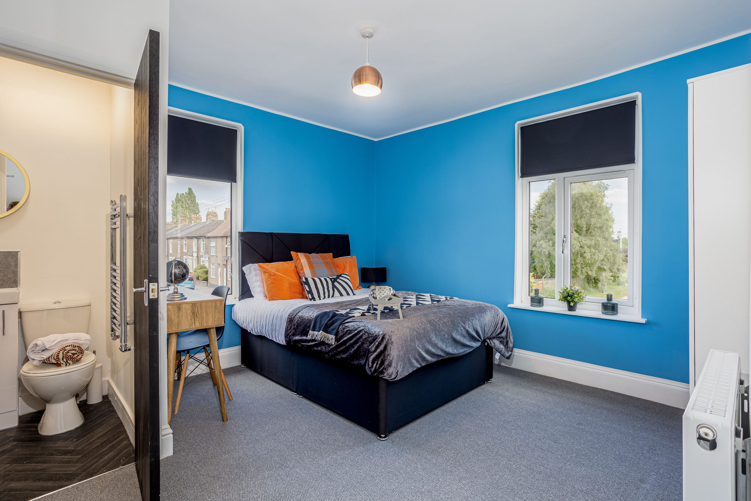 28d8aacb-06c9-41bb-b01a-0ecad146d55cjpg | RENT A ROOM | Professional House Shares in Warwickshire & West Midlands Bright blue-walled bedroom with a black bed frame, orange cushions, and two windows with dark roller blinds; a small bathroom is visible to the left.