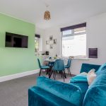 3376c68e-cc3d-459d-ab5e-eb5cba6ea0fajpg | RENT A ROOM | Professional House Shares in Warwickshire & West Midlands Living room with a green feature wall and wall-mounted TV, round dining table for four, blue chairs, and a hanging plant display in the corner.