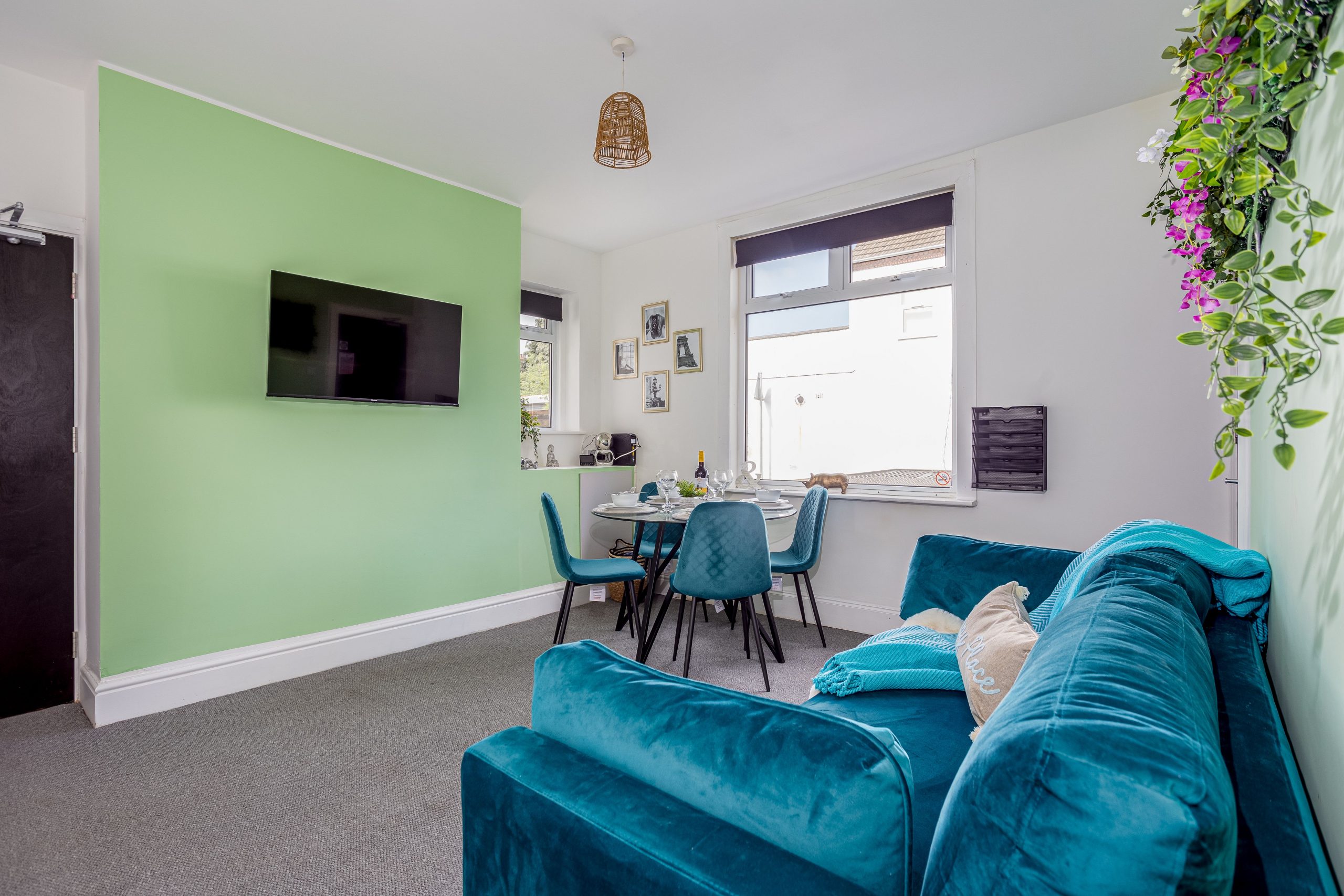 3376c68e-cc3d-459d-ab5e-eb5cba6ea0fajpg | RENT A ROOM | Professional House Shares in Warwickshire & West Midlands Living room with a green feature wall and wall-mounted TV, round dining table for four, blue chairs, and a hanging plant display in the corner.