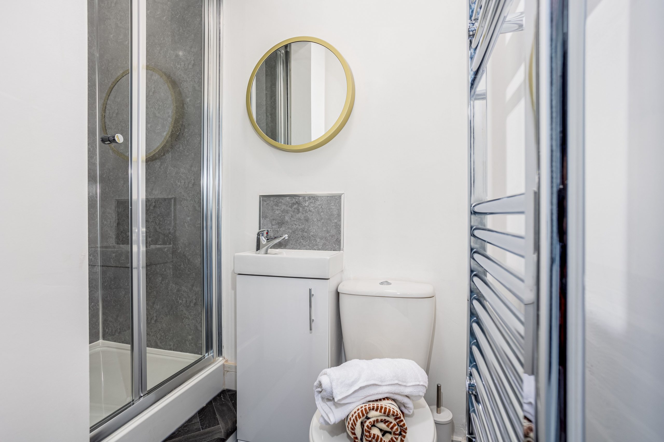 4cb0e074-8167-470b-823e-e0d1167bce66jpg | RENT A ROOM | Professional House Shares in Warwickshire & West Midlands Compact modern bathroom with a glass shower enclosure, round gold-framed mirror, white sink and toilet, and towels on the toilet.