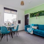 a3550fe7-0a27-40be-8ee1-9588967b8dd7jpg | RENT A ROOM | Professional House Shares in Warwickshire & West Midlands Cozy dining area with turquoise chairs around a glass table set for a meal; teal sofa and green wall with hanging greenery in the background.