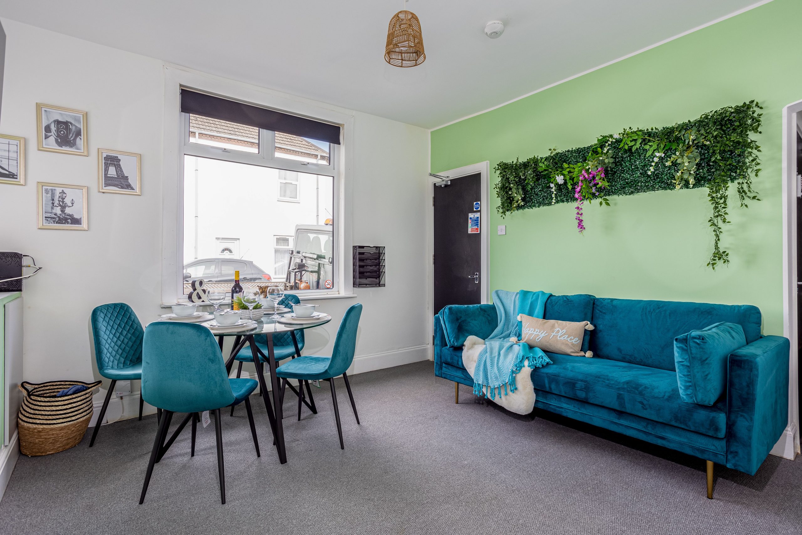 a3550fe7-0a27-40be-8ee1-9588967b8dd7jpg | RENT A ROOM | Professional House Shares in Warwickshire & West Midlands Cozy dining area with turquoise chairs around a glass table set for a meal; teal sofa and green wall with hanging greenery in the background.