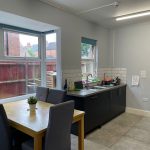 Bright dining area with a wooden table and gray cushioned chairs, overlooking a kitchen with a double sink and large windows.