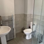 Compact modern bathroom with a white pedestal sink, chrome towel radiator, toilet, and a glass corner shower.