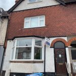 Red brick terraced house with a white bay window on the ground floor and a for-rent sign by the front door; several wheelie bins outside.