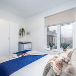 A bright, minimalist bedroom with a white wardrobe, small shelves, and a bed covered in a blue quilt and patterned pillows next to a large window.