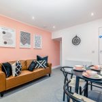 Cozy living area with a peach accent wall, three Bauhaus posters above a tan leather sofa, and a round dining table set for four with black chairs.
